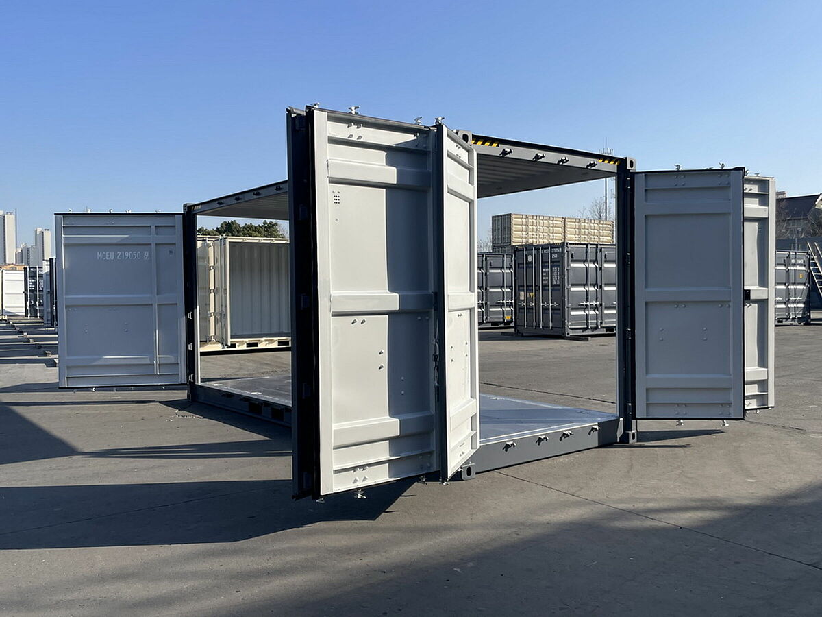 Ein geöffneter 20-Fuß-High-Cube-Container mit All-Side-Doors steht auf einem Containerplatz. Die Seiten- und Stirnwände sind vollständig aufgeklappt und geben den Blick auf den Innenraum frei. Der Container ist hellgrau lackiert, steht auf einem asphaltierten Untergrund und ist von weiteren gestapelten Containern umgeben. Im Hintergrund sind Bäume, Industriegebäude und ein klarer blauer Himmel zu sehen.