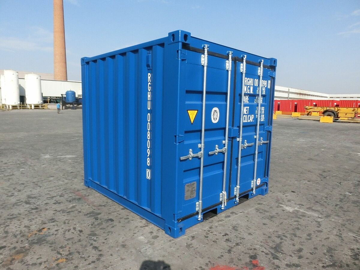 Ein blauer 8-Fuß-Minikontainer steht auf einer betonierten Freifläche eines Industriegeländes. Der Container hat stabile Verriegelungsstangen an der Tür und ist mit weißen Identifikationsnummern sowie Warnsymbolen beschriftet. Im Hintergrund sind weiße Tanks, ein Schornstein und weitere Container zu sehen.