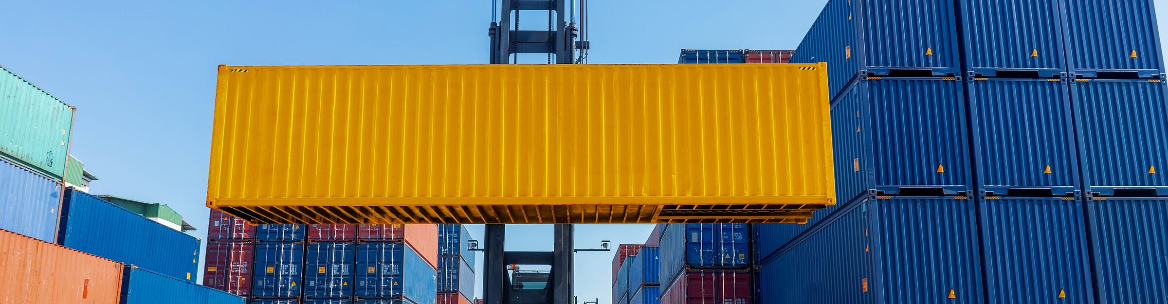 Gelber Frachtcontainer wird per Kran zwischen gestapelten Containern verladen.