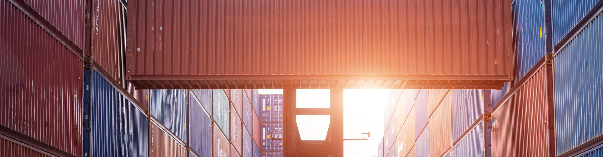 Frachtcontainer wird zwischen gestapelten Containern bei Sonnenuntergang verladen.
