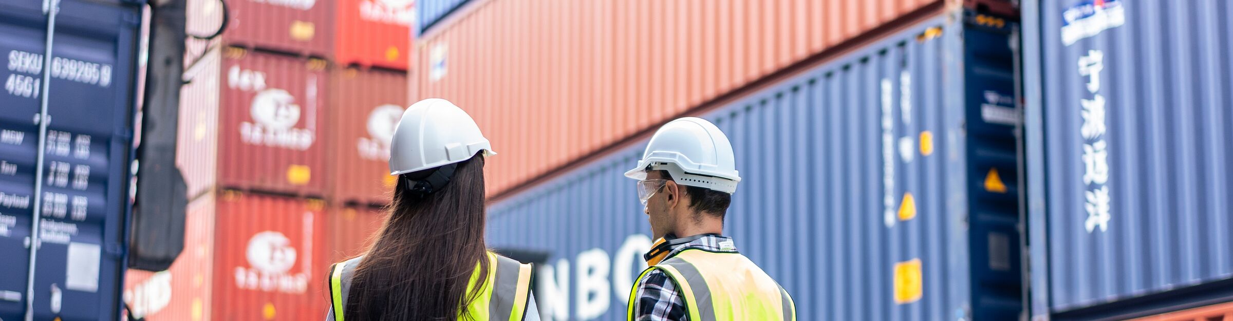 Zwei Logistikmitarbeitende mit Schutzhelm und Warnweste prüfen Container im Frachtbereich.