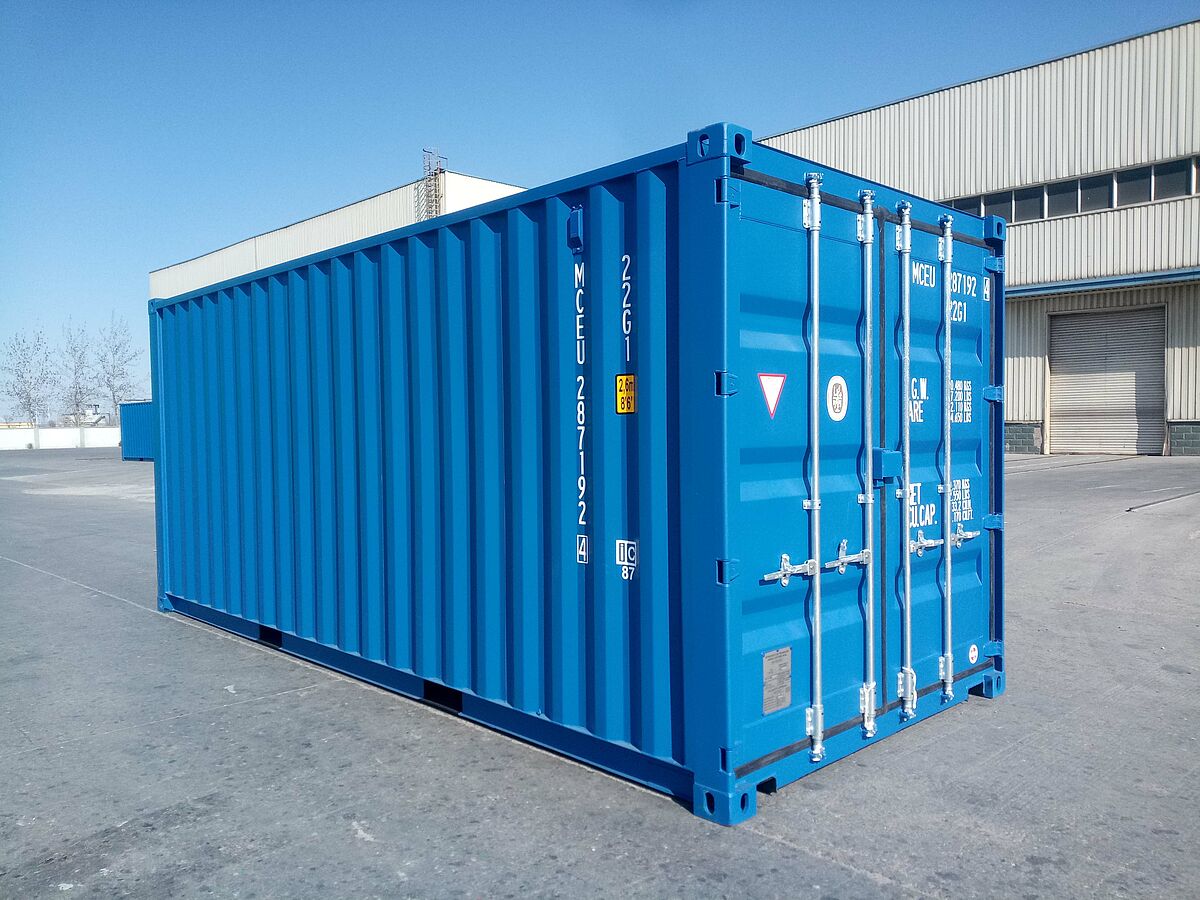 Ein blauer 20-Fuß-Standard-Seecontainer steht auf einer asphaltierten Freifläche vor einer Industriehalle. Der Container besitzt verstärkte Ecken, Verriegelungsstangen an der Tür und ist mit verschiedenen technischen Kennzeichnungen und Nummern beschriftet.